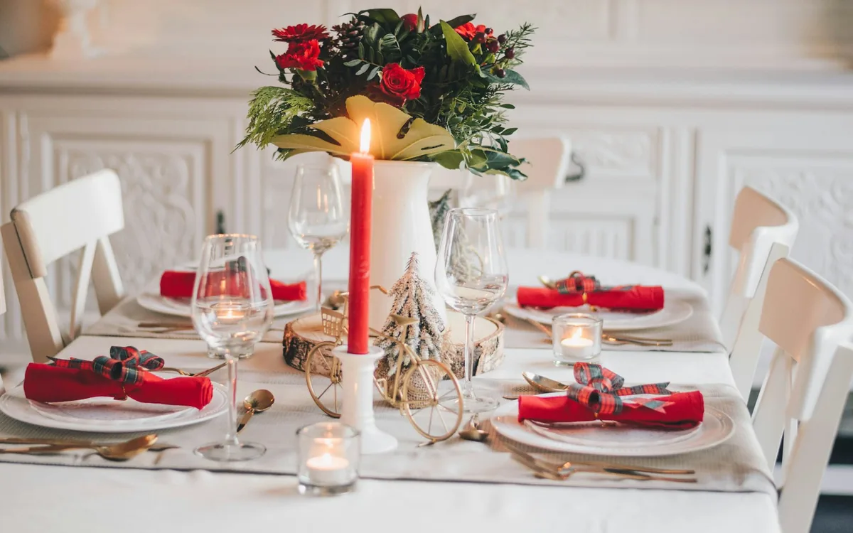 A holiday table laid in white and red: candle, greenery and set places.