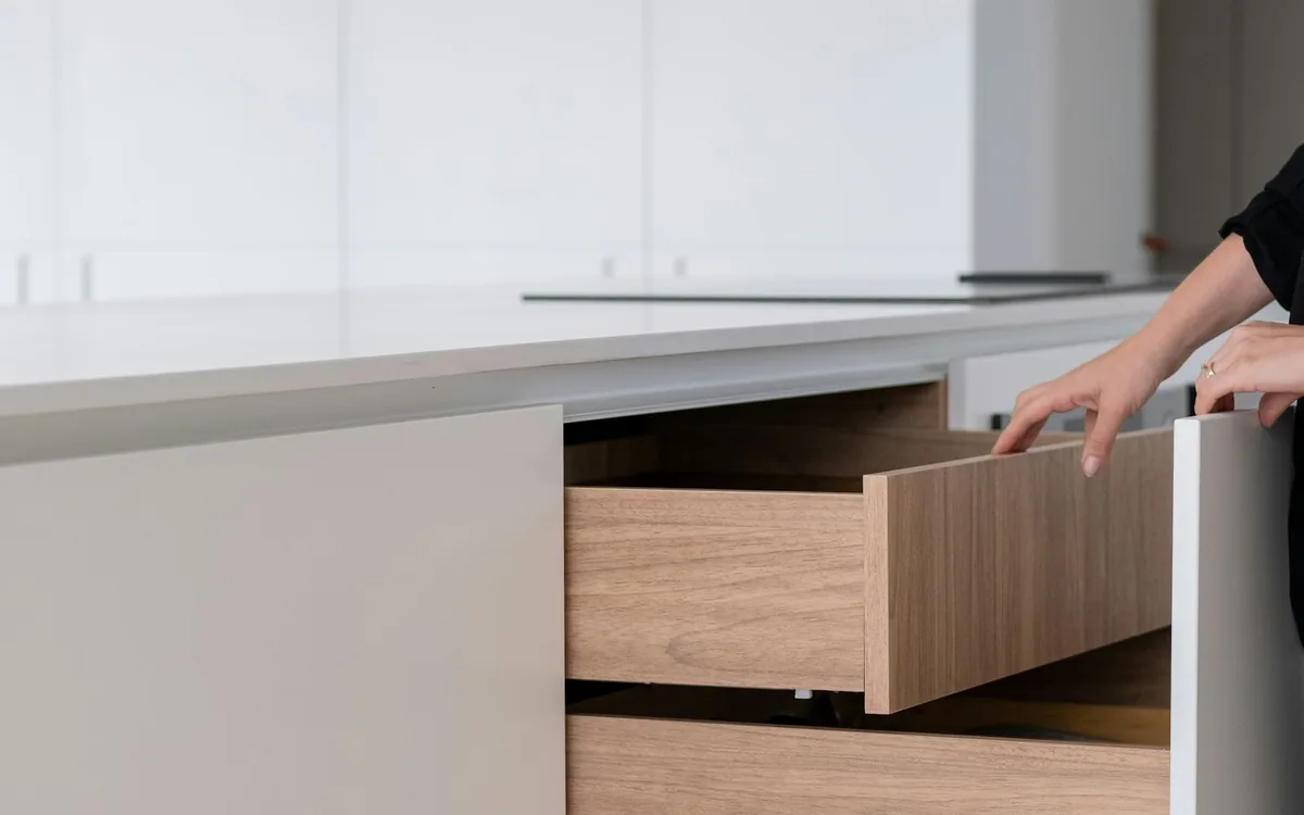 A hand opening a timber-lined drawer in a handleless white kitchen.