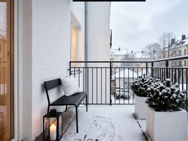 Minimalist balcony in winter with a black metal bench, two white cube planters of dwarf evergreens dusted with snow and one glowing black lantern