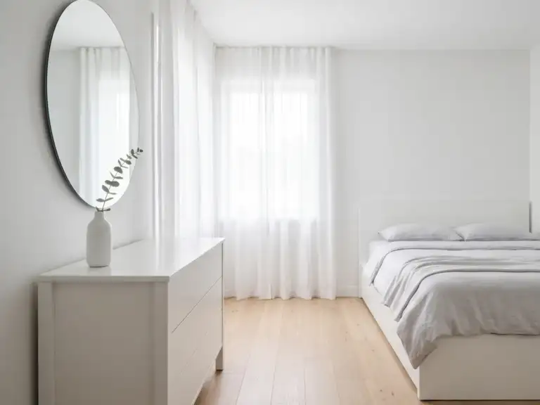 Small minimalist white bedroom with a large frameless round mirror above a slim white dresser, a low bed with pale gray bedding and a single vase
