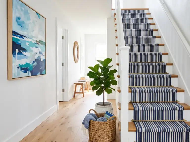 A narrow staircase with white risers, honey oak treads and a navy and white striped flat-weave runner with brass rods, beside a blue abstract painting on a white wall.
