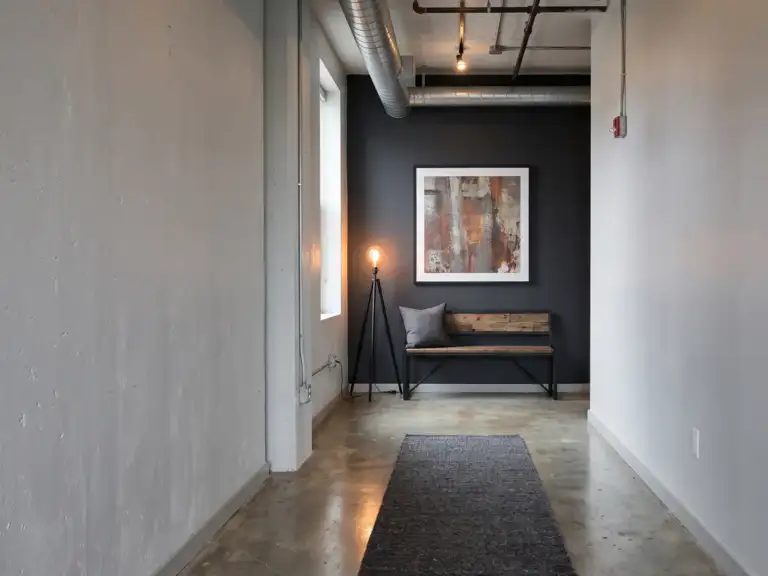 Industrial hallway with light gray walls, a charcoal end wall, a black metal bench, a tripod floor lamp with a bare bulb and a large framed abstract print.